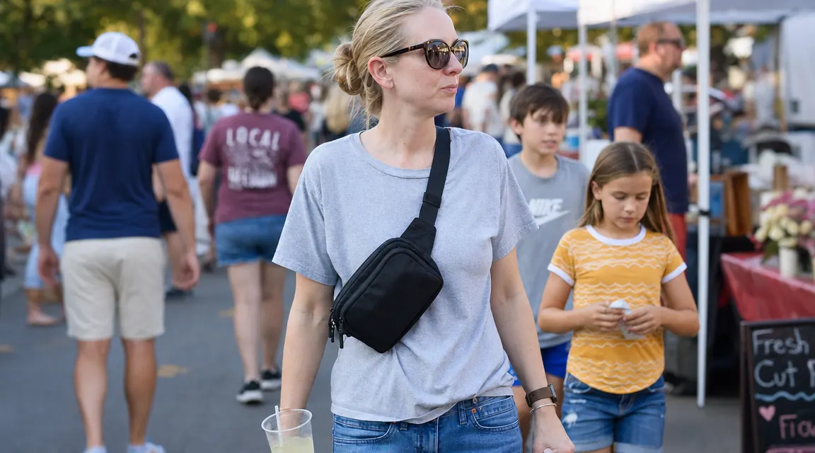 Woman wearing a black crossbody sling bag over her shoulder at a busy outdoor market, kids visible nearby