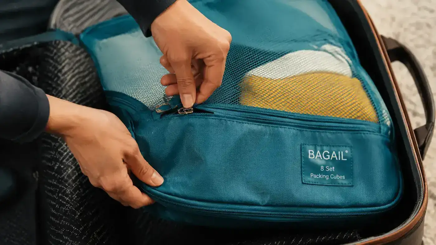 Close-up of hands unzipping a BAGAIL packing cube, showing the double zipper pull and mesh top panel